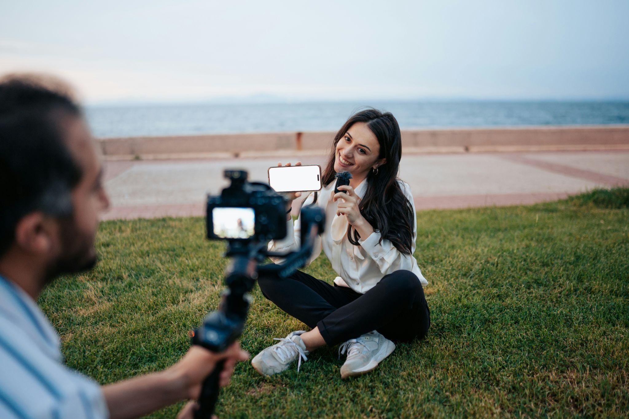 Creator sitting on grass near the water holding a phone and microphone while a videographer records her for social media clips and repurposed posts.
