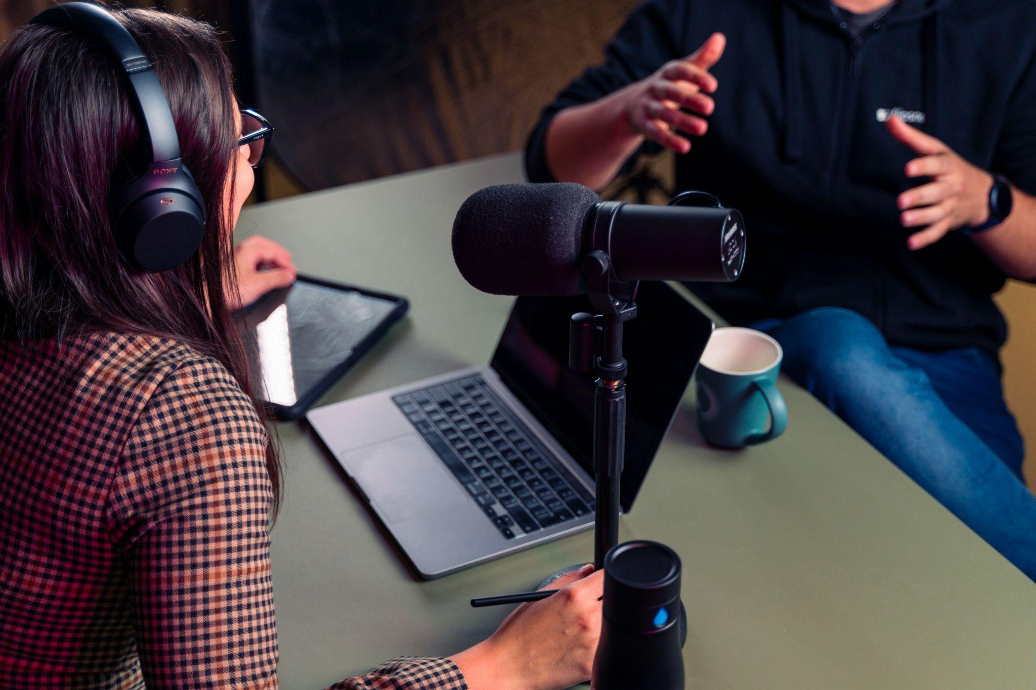 Two marketers recording a podcast conversation with headphones, laptop, and microphone to capture ideas for later repurposed content.