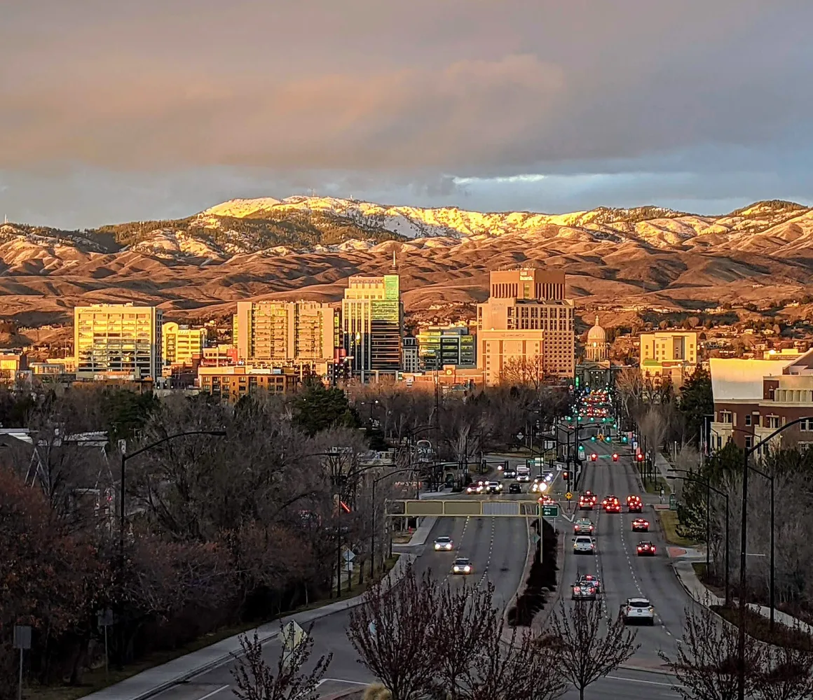 Boise's downtown and small capitol dome at sunset, snow-tipped foothills behind a busy boulevard