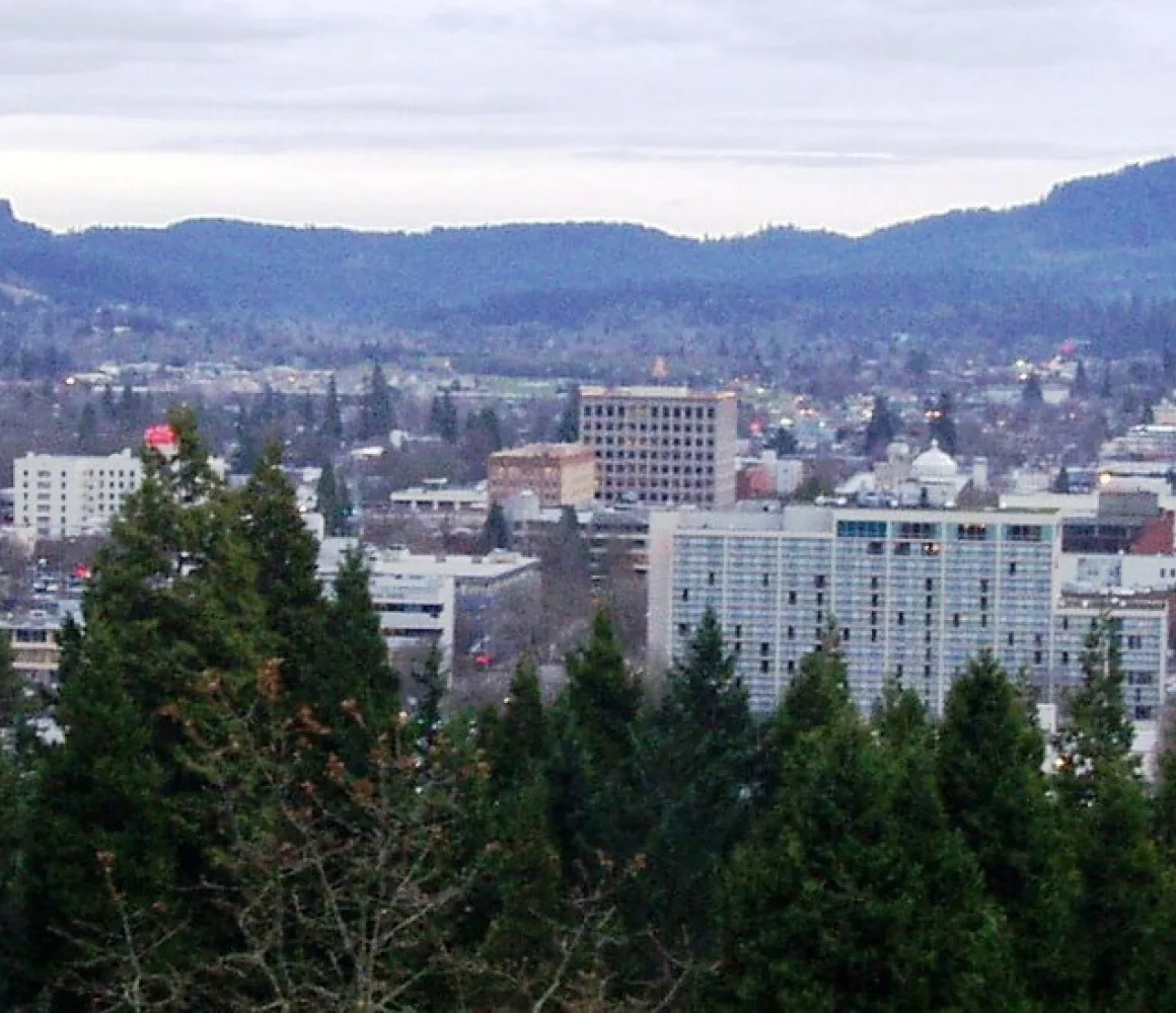 Eugene seen over evergreen treetops from a hillside, with forested buttes under grey cloud