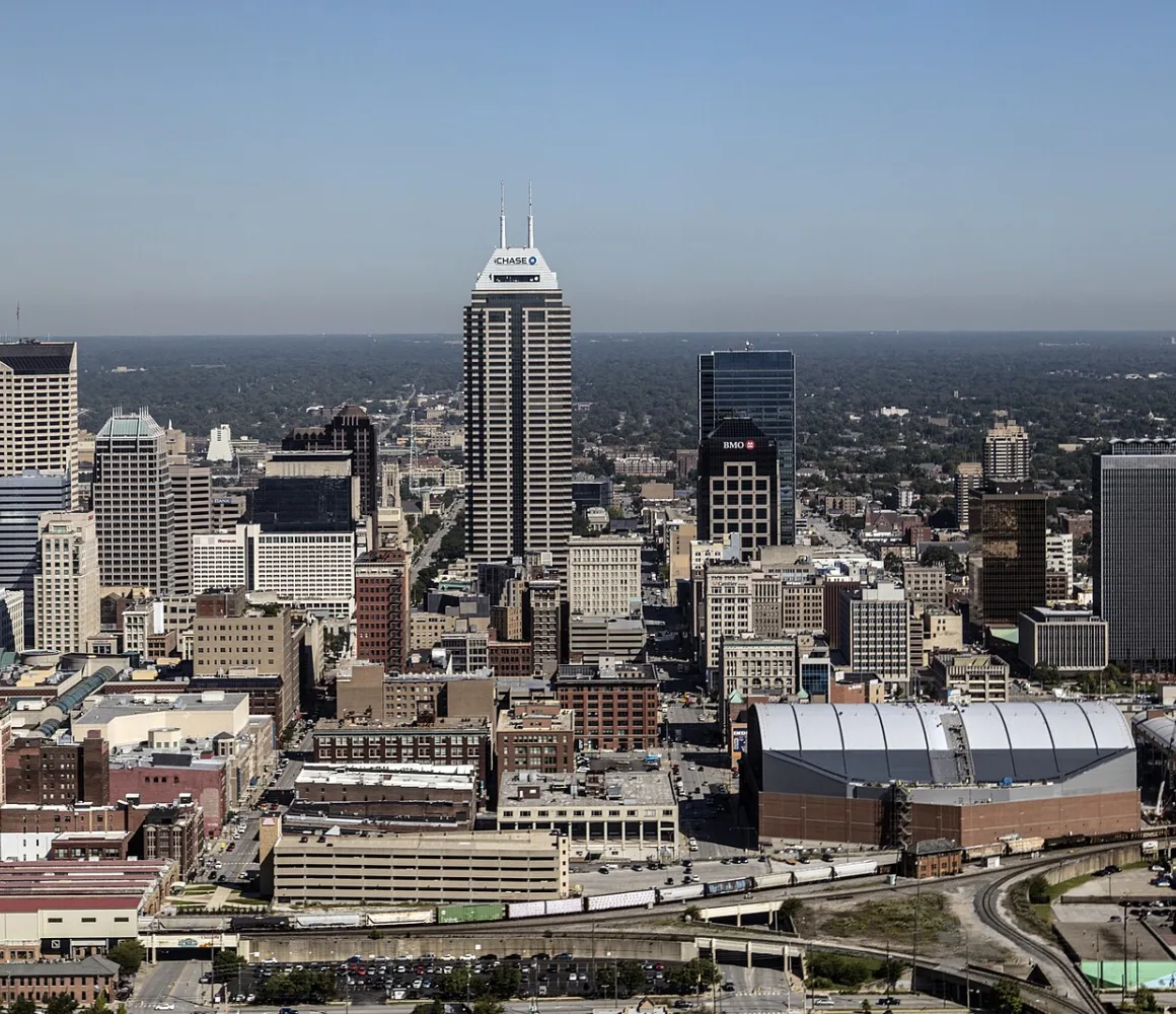 Aerial view of downtown Indianapolis, a spire-topped tower and arched-roof arena under hazy sky
