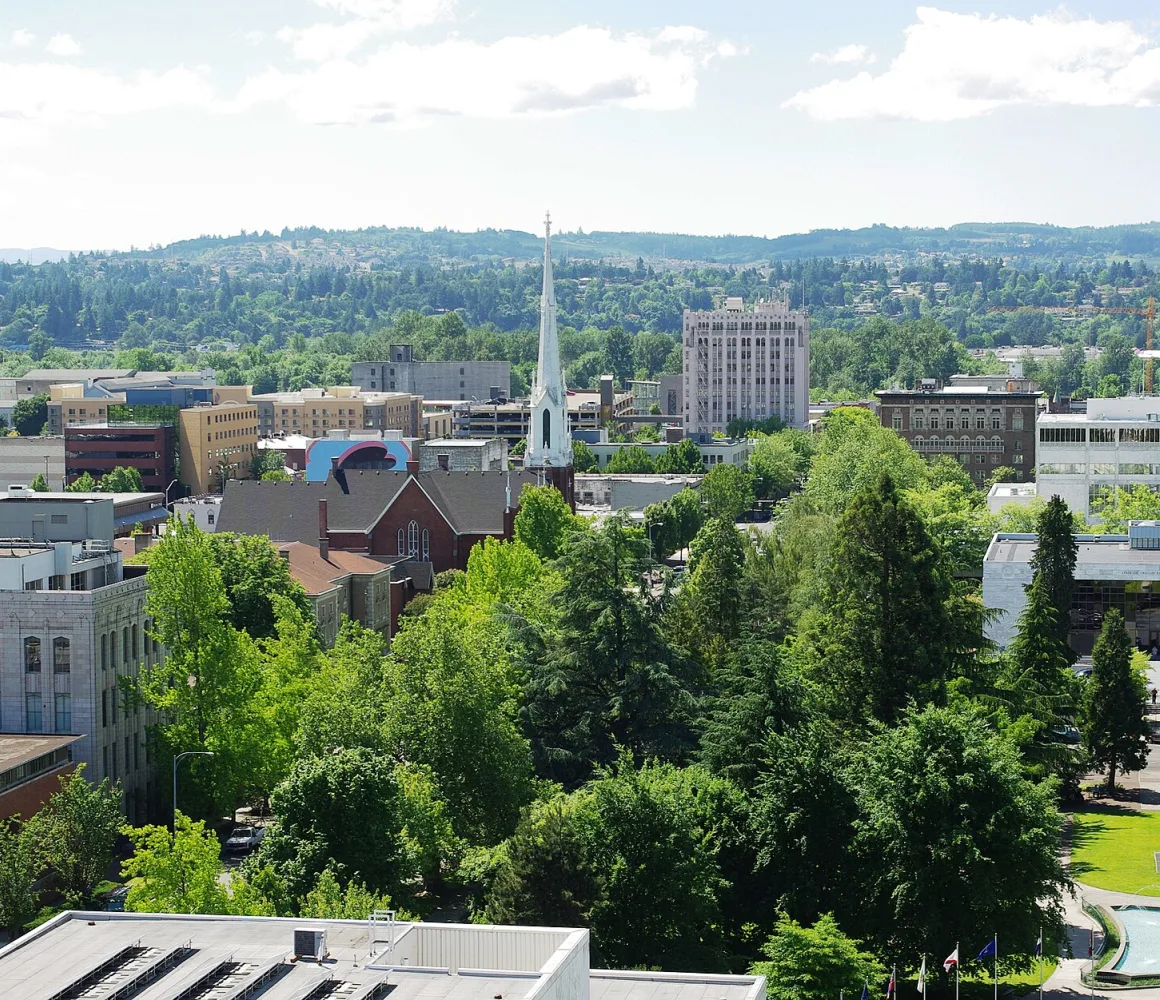 A white church steeple above leafy downtown Salem, with forested hills beyond and a fountain in a park