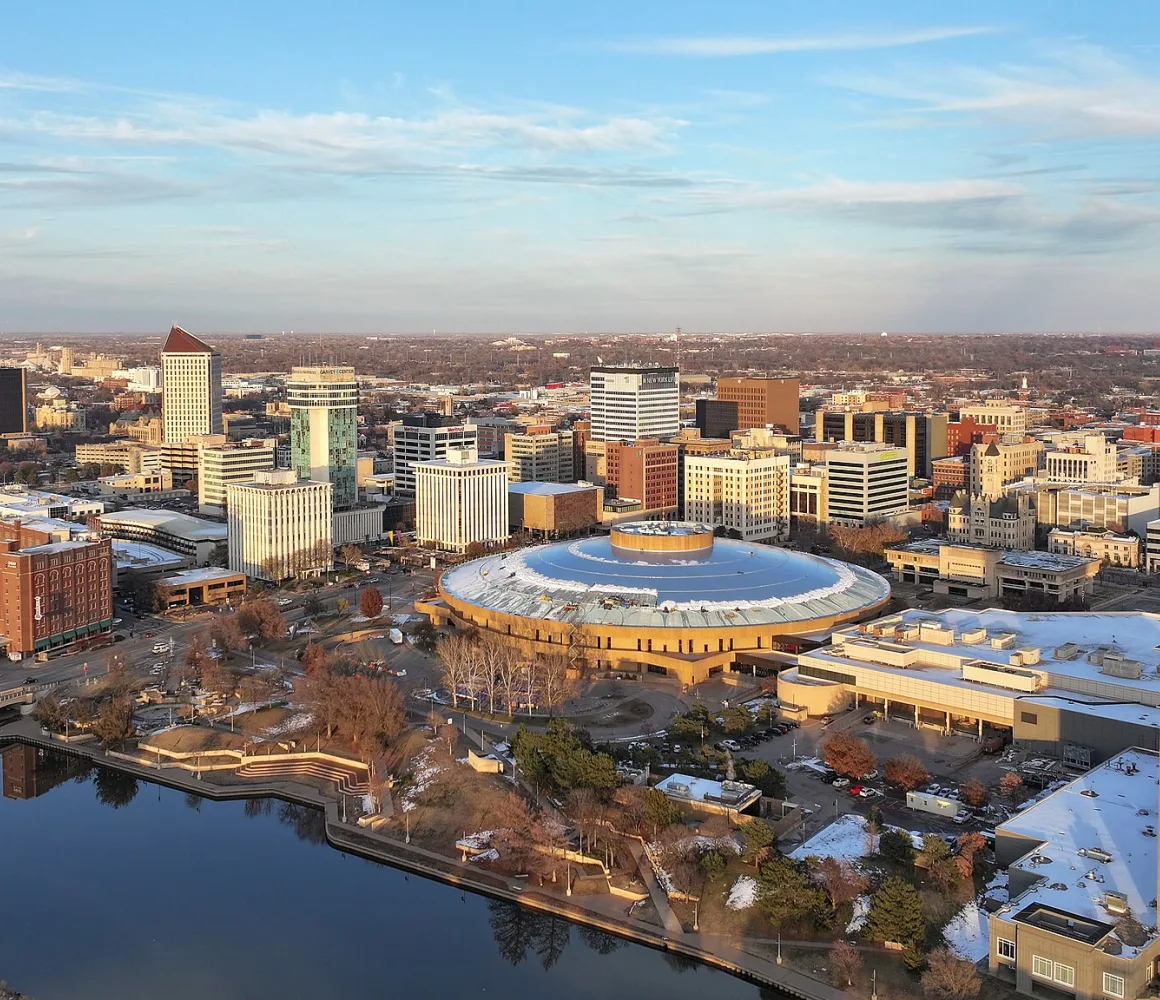 Snow-dusted aerial of Wichita at golden hour, a round arena beside the river and low downtown skyline
